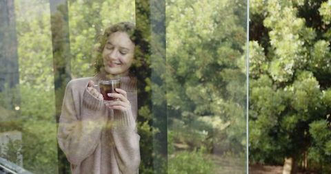 Woman in Cozy Sweater Enjoying Tea Behind Window with Nature View