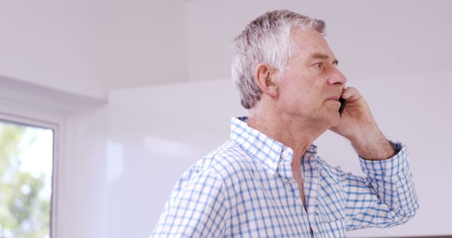 Senior man with gray hair talking on phone in casual wear and using smartphone in a bright room. This can be used to depict concepts like staying connected, using technology, communication, senior lifestyle, or indoor activities.