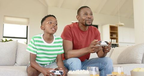 African american father and son playing video games on couch sharing popcorn and bonding