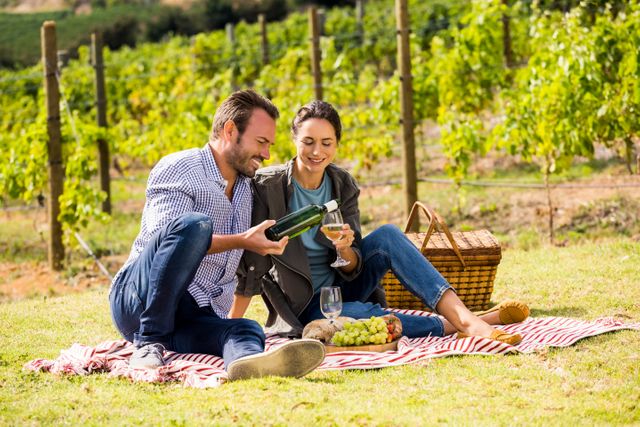 Couple sitting on blanket in vineyard, man pouring wine into glass held by woman, grapes and picnic basket beside them. This image is perfect for themes related to romance, outdoor activities, relaxation, vineyards, wine tastings, and couple getaways.