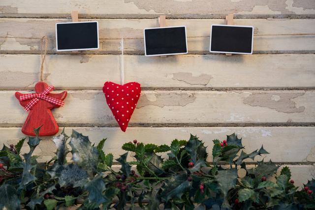 Festive Christmas decorations hanging on rustic wooden wall include small red ornaments and chalkboards. Holly leaves and berries at bottom add to holiday cheer. Ideal for use in holiday greeting cards, Christmas craft ideas, festive social media posts, and seasonal home decor inspiration.