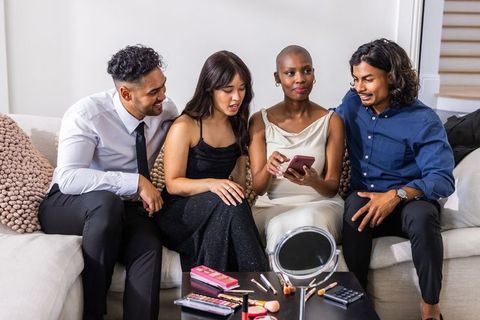 Diverse friends sharing makeup palettes and smartphone on sofa during party prep