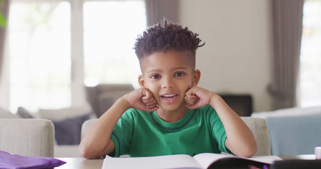 Young boy in a green shirt smiling while sitting at table with an open book. Ideal for educational content, family lifestyle blogs, and homeschooling resources.