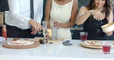Diverse friends grating cheese and assembling homemade pizzas at kitchen island party
