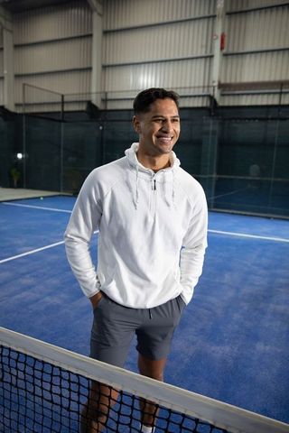 Smiling male athlete standing courtside on indoor blue tennis court wearing white hoodie and shorts