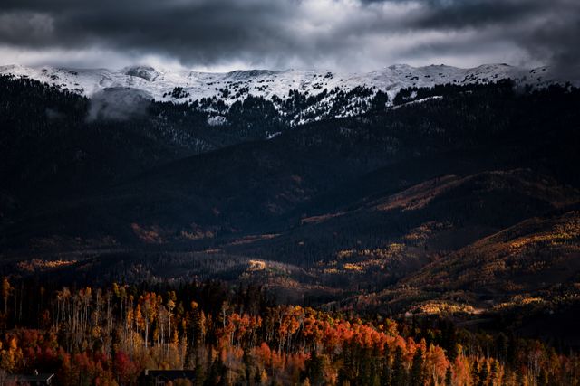 Snow-capped mountains rise above a mixed forest transitioning to autumn colors, with vibrant oranges and reds juxtaposed against dark evergreen trees. Looming dark clouds above create a striking, moody atmosphere. This scenic view is ideal for nature and travel blogs, outdoor adventure promotions, seasonal calendars, and postcards. It captures the essence of changing seasons in a pristine wilderness setting.