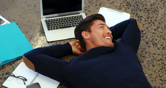 A young Caucasian man lies on the floor with his hands behind his head, surrounded by work materials and a laptop, with copy space. His relaxed posture and smile suggest a break from work or a moment of satisfaction after completing a task.