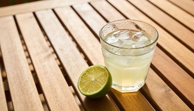 Sunlight catching faceted glass of iced limeade with halved lime on wooden slatted table