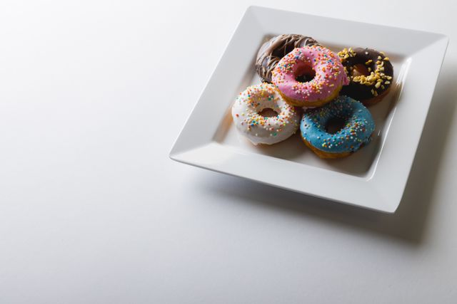 Assorted colorful donuts with various toppings on a white square plate. Ideal for use in food blogs, dessert menus, bakery advertisements, and social media posts promoting sweet treats and indulgent snacks.