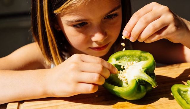Young girl removing seeds from green bell pepper on wooden board, kitchen learning moment
