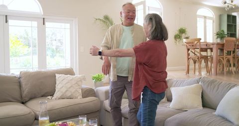 Mature asian couple dancing and reaching in bright modern living room with smartwatch and snacks