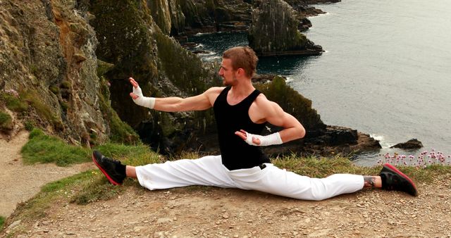 A young Caucasian male athlete performs a split on a cliff overlooking the sea, with copy space. His focus and balance showcase a blend of strength and flexibility in a breathtaking natural environment.