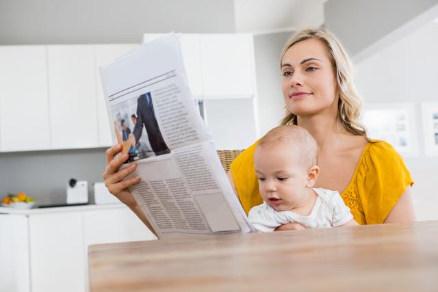 Mother reading a newspaper while holding her baby boy in a modern kitchen. Ideal for topics on parenting, family life, motherhood, and daily routines. Can be used in articles, blog posts, and advertisements focusing on work-life balance, family activities, or home living.
