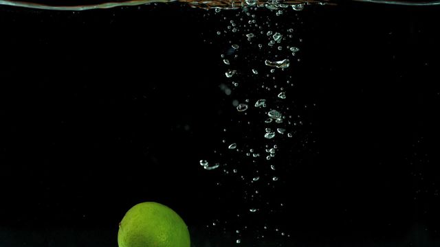 Green lime dropping into water, creating bubbles on black background. Useful for topics related to freshness, underwater photography, health, hydration, citrus fruit promotion, advertisement. Ideal for food and beverage marketing materials, water and fruit-related advertisements, and healthy lifestyle blogs.