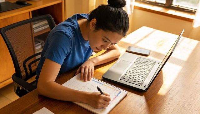 Asian woman writing in planner while working at home desk with laptop and smartphone