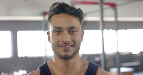 Young adult male fitness trainer with short dark hair and a beard enjoys positive moment in gym environment. Dressed in black tank top, showing fit and strong physique, sets motivational example for workout enthusiasts. Clear view of gym equipment in background further enhances focus on gym setting. Perfect for promoting health and fitness products, personal training services, or active lifestyle campaigns.