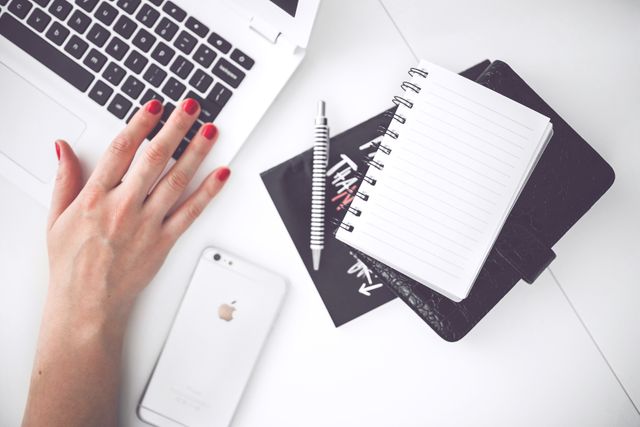 Woman's hand with red nails using laptop while notepad and smartphone lying on desk. Useful for content about productivity, work from home, modern workspace, business applications, and office supplies.