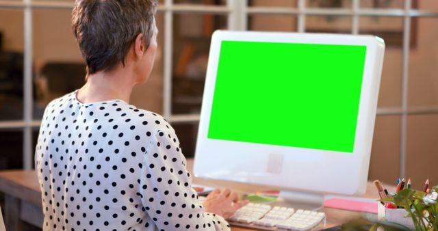 Senior woman working on desktop computer with green screen in modern office environment. Useful for demonstrating software, website display, or computer applications. Ideal for marketing, business presentations, and technology-oriented content.