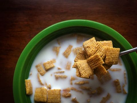 Spoon scooping whole grain square cereal from green bowl with milk on wooden table