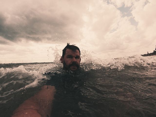 Man swimming in rough ocean waves on a cloudy day, surrounded by splashing water. The dark, overcast sky suggests stormy weather, adding a dramatic effect to the scene. Useful for themes of adventure, nature, resilience, and outdoor activities. Can be used in blogs about swimming, ocean adventures, and coping with weather challenges in coastal areas.