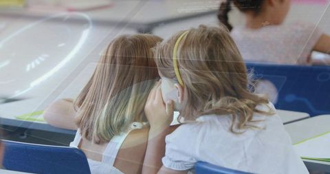 Two blonde girls whispering at classroom desks sharing secret during primary school lesson