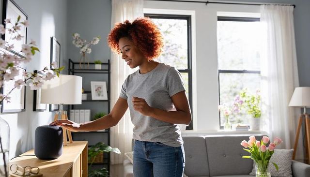 Young woman controlling smart speaker with touch in modern sunlit living room interior