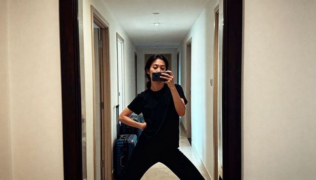 Asian woman taking mirror selfie in hotel hallway with suitcase and casual outfit