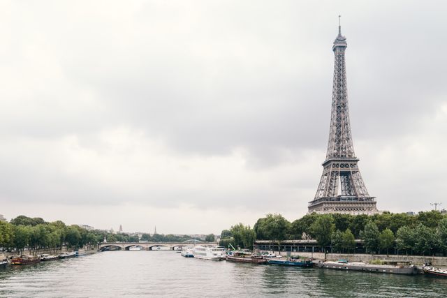 The scene captures the Eiffel Tower standing tall on an overcast day, with the tranquil Seine River flowing in the foreground. The iconic Parisian landmark is enveloped by lush green trees, and several boats line the riverbank. This visual can be ideal for travel brochures, tourism websites, educational materials about French culture, historic sites, or desktop wallpapers evoking a sense of picturesque serenity.