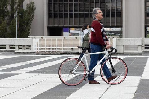 Senior man walking white bicycle across urban plaza with asian woman passing by, city lifestyle