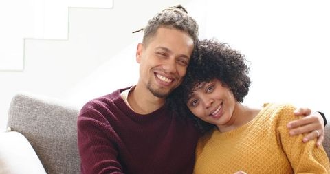 Smiling young couple cuddling on sofa in sunlit living room wearing cozy sweaters