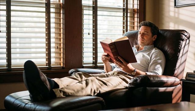 Man reading hardcover while reclining on leather recliner by sunlit wooden blinds