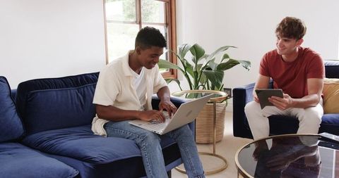Diverse young men collaborating on laptop and tablet in cozy blue velvet living room