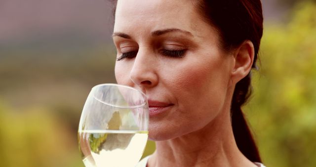Woman calmly enjoying a glass of white wine outdoors. This can be used for any lifestyle and leisure content, wine tasting events, vineyard promotions, relaxing and laid-back concepts. Ideal for advertisements promoting wine, leisure time activities, or serene outdoor experiences.