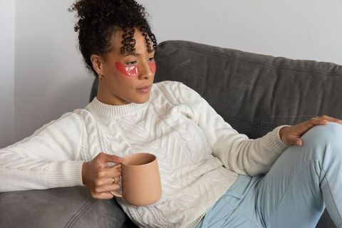 African american woman lounging on sofa wearing red hydrogel under-eye patches holding mug