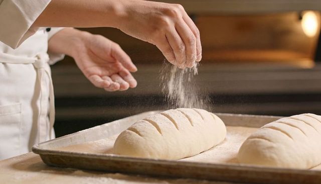Baker hands sprinkling flour over scored artisan bread loaves on floured baking tray
