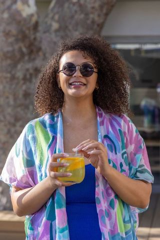 African american woman smiling and holding citrus cocktail on sunny patio in pastel kimono