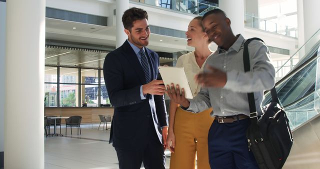 Three business professionals standing together in a modern office lobby, engaging in a lively discussion while looking at a tablet. Ideal for depicting teamwork, collaboration, corporate culture, modern workspaces, and professional communication in marketing materials or corporate presentations.