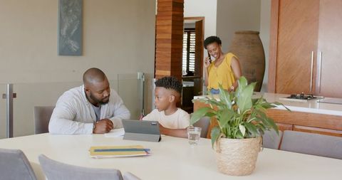 African american father helping son with tablet, mother on phone at modern dining table