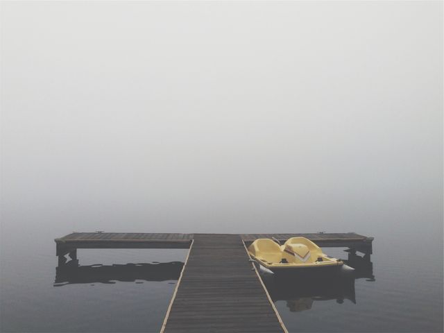 Calm volerec setting showcasing wooden dock leading to a misty lake with two yellow paddle boats. Perfect for themes like tranquility, nature's serenity, outdoor adventures, or peaceful escapes. Ideal for travel brochures, blog posts about relaxation or nature, and background images conveying calmness.