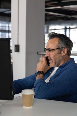 Middle-aged professional talking on phone at desk with coffee in open-plan office