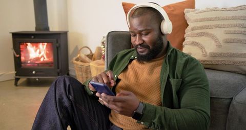 African american man relaxing by fireplace wearing headphones scrolling smartphone on sofa
