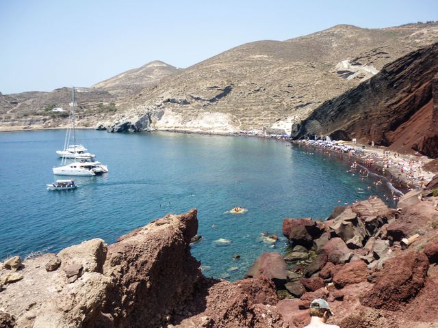 Tourists enjoying sunny day at the famous Red Beach in Santorini, Greece, with boats anchored in clear blue waters. Perfect for promoting travel destinations, vacation packages, or Mediterranean cruises. Ideal for blog posts, travel guides, or marketing material highlighting exotic locations.