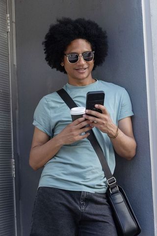 Man leaning in doorway checking phone while holding coffee and messenger bag