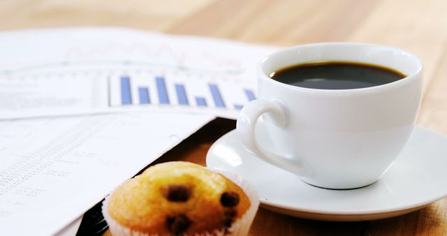 A cup of coffee sits next to a muffin and financial documents with graphs, indicating a business breakfast or a work break. The setting suggests a professional reviewing performance metrics or preparing for a financial meeting, with copy space.