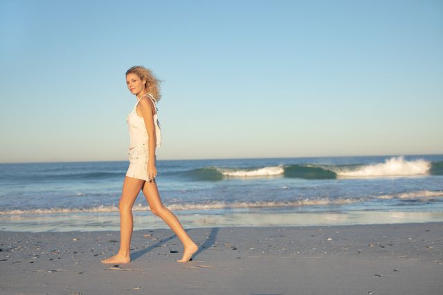 Woman walking barefoot on beach at sunset, enjoying peaceful and serene environment. Ideal for use in travel brochures, wellness blogs, lifestyle magazines, and advertisements promoting relaxation and vacation destinations.