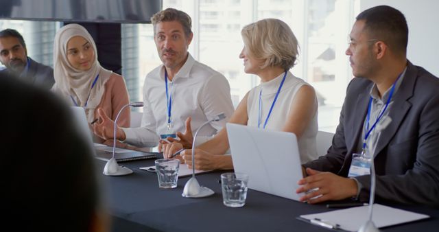Front view of a young Caucasian businessman sitting at the centre of a row of five diverse business delegates at a table with microphones in front of them at a business conference. The young man is speaking and looking to the audience while his colleagues on the panel have turned towards him to listen