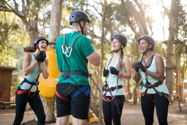 This image shows a coach interacting with a group of trainees in a forest adventure park. They are all wearing protective helmets and safety gear, indicating they are about to engage in an outdoor activity. This image can be used for promoting team-building exercises, adventure parks, outdoor training programs, and fitness activities. It highlights themes of teamwork, motivation, and fun in a natural setting.
