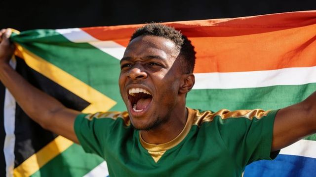 Triumphant south african football fan cheering with flag and green jersey in stadium