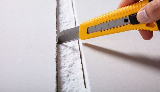 Hand holding yellow utility knife scoring drywall joint releasing gypsum dust closeup