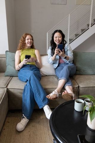 Diverse friends relaxing on sofa, laughing and using smartphones in cozy living room at home
